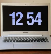 Silver laptop on a wooden desk showing a digital clock at 12:54.